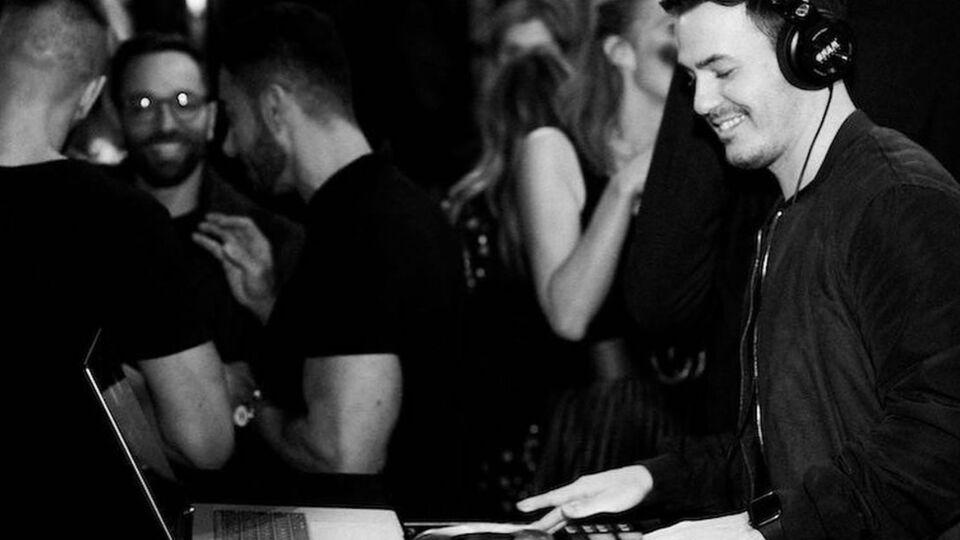 DJ Corey in headphones smiling at the decks beside dancing guests, in black and white