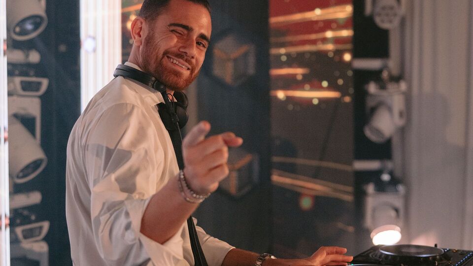 DJ Nathan smiling and pointing from the DJ booth, moving-head lights behind him