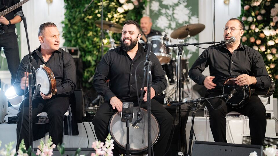 Three percussionists in black shirts playing frame drums on a stage with a flower wall
