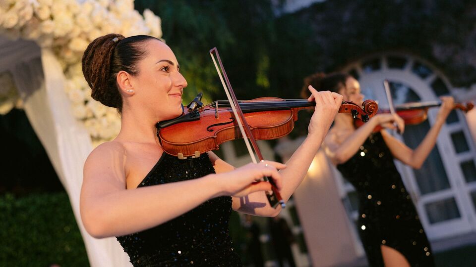 Two violinists in black sequined gowns playing beside a white floral ceremony arch at dusk