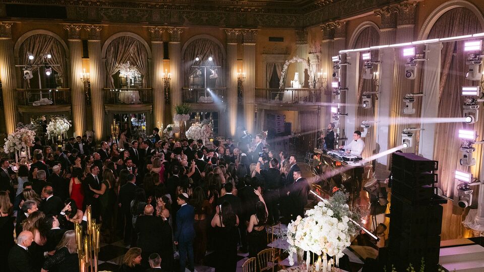 Crowded ballroom with gilded columns and arched windows, guests dancing by the DJ stage