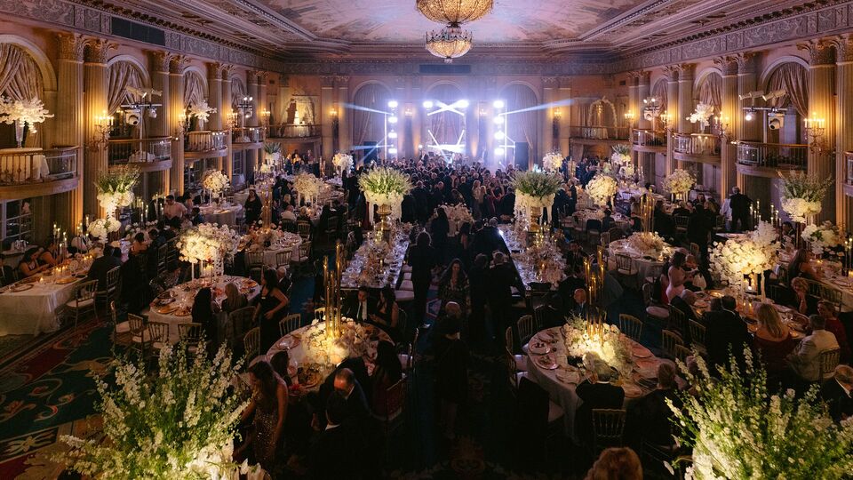 Historic ballroom with a painted ceiling and chandeliers, guests at candlelit tables with tall white flowers