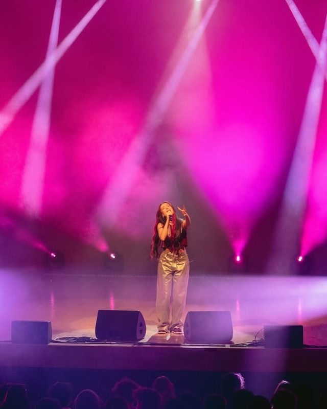 A singer alone on a large stage under pink haze and crossing beams