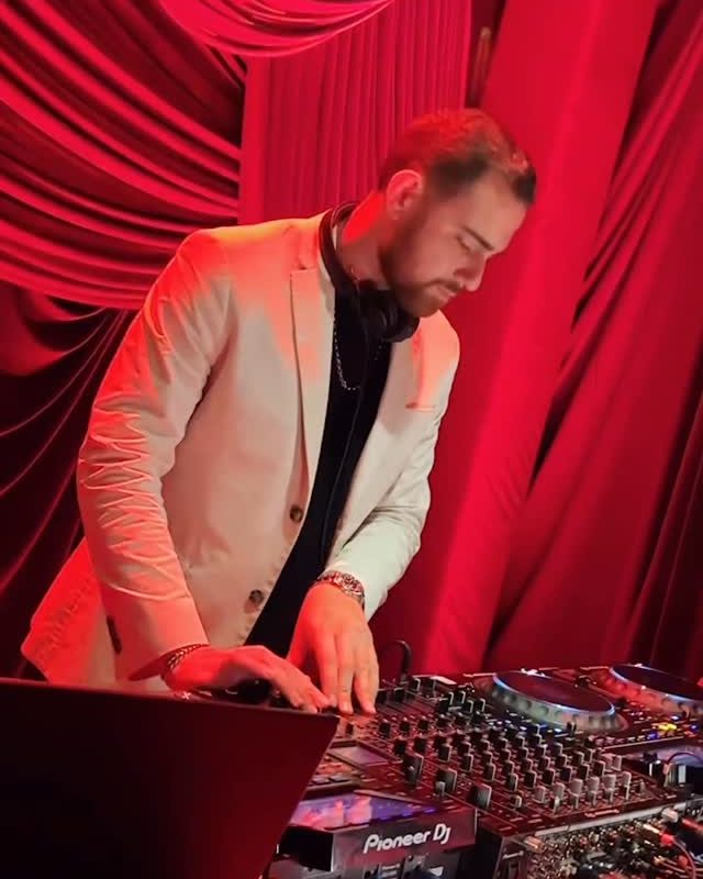 A DJ in a white jacket at the decks in a room hung with red drapes