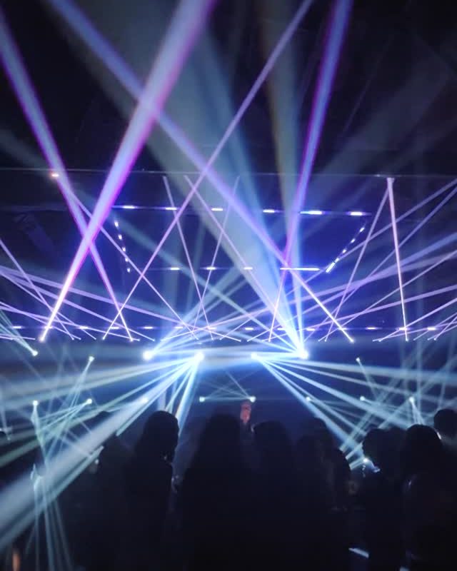 White and violet beam lights crossing above a crowd in a dark warehouse