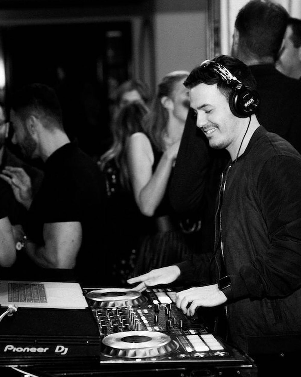 DJ Corey in headphones at a Pioneer controller with guests behind him