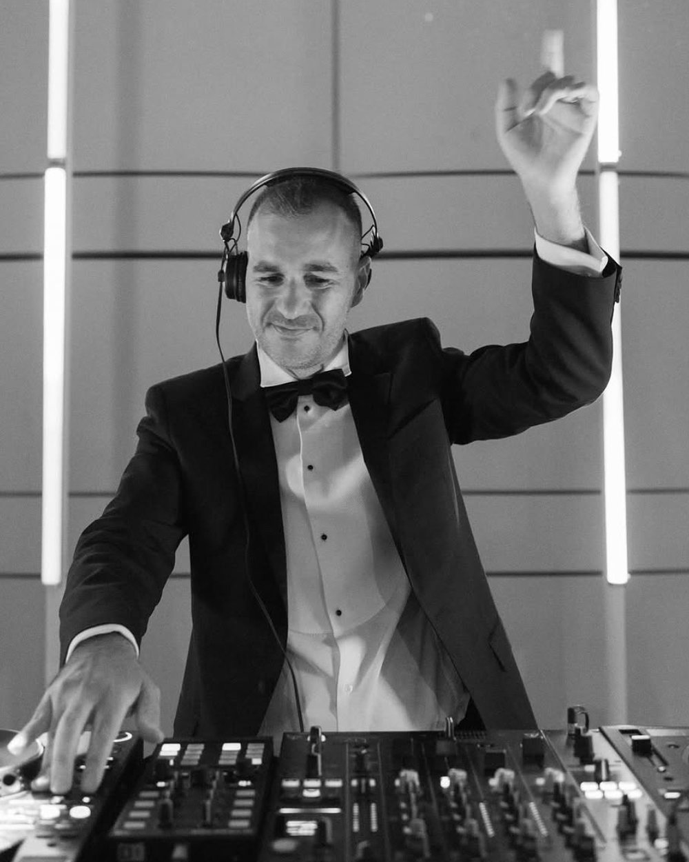 DJ Oren in a tuxedo and headphones with one hand raised over the mixer, in black and white