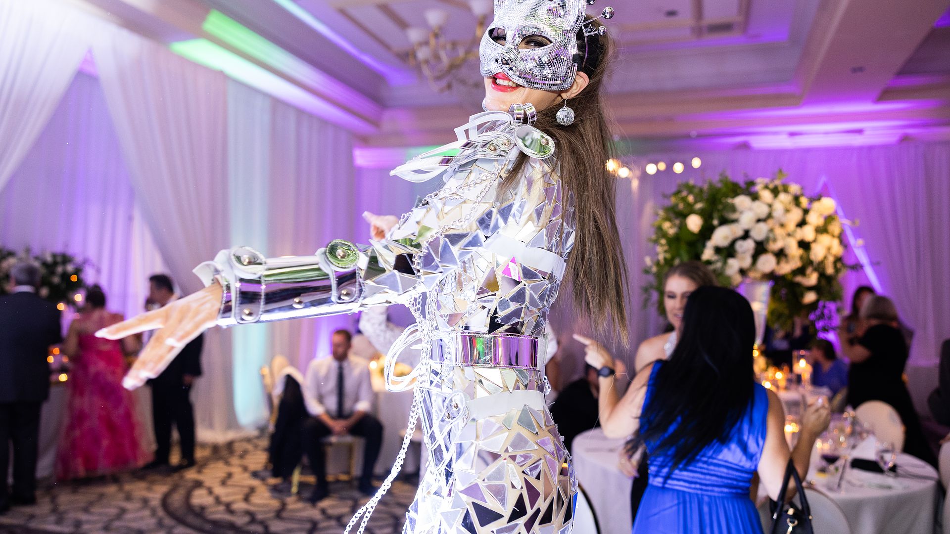 A performer in a full mirror-mosaic costume and jewelled mask moving among seated guests at a ballroom reception