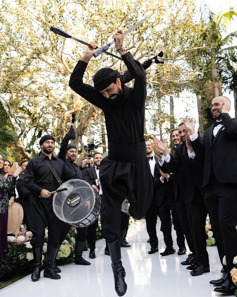 A dancer leaping mid-air with two raised swords during a wedding procession, guests applauding