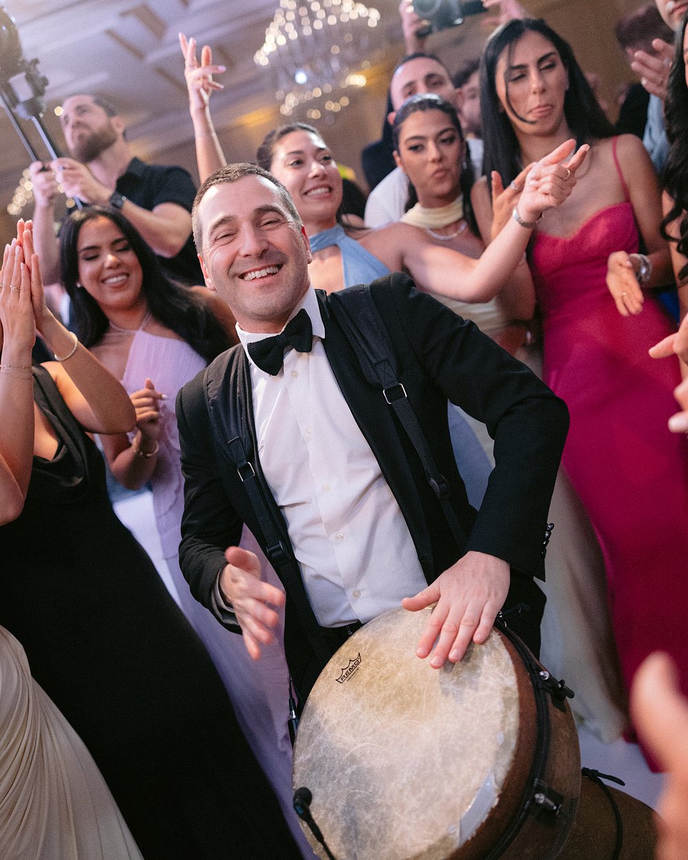 A percussionist playing a frame drum surrounded by guests dancing at a reception
