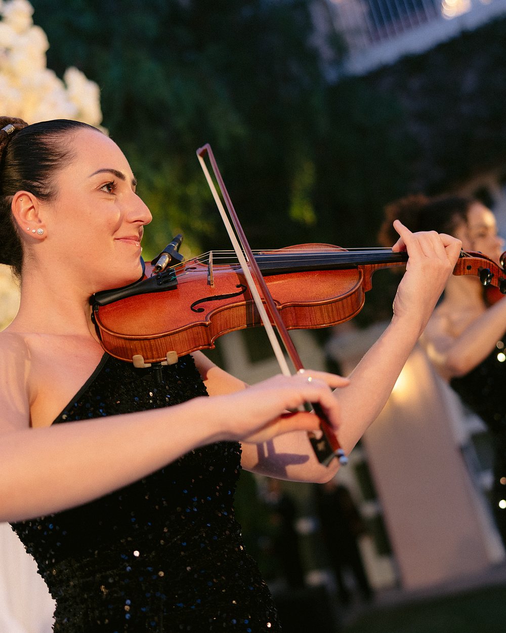 A violinist playing an acoustic violin outdoors at dusk, a second player behind her