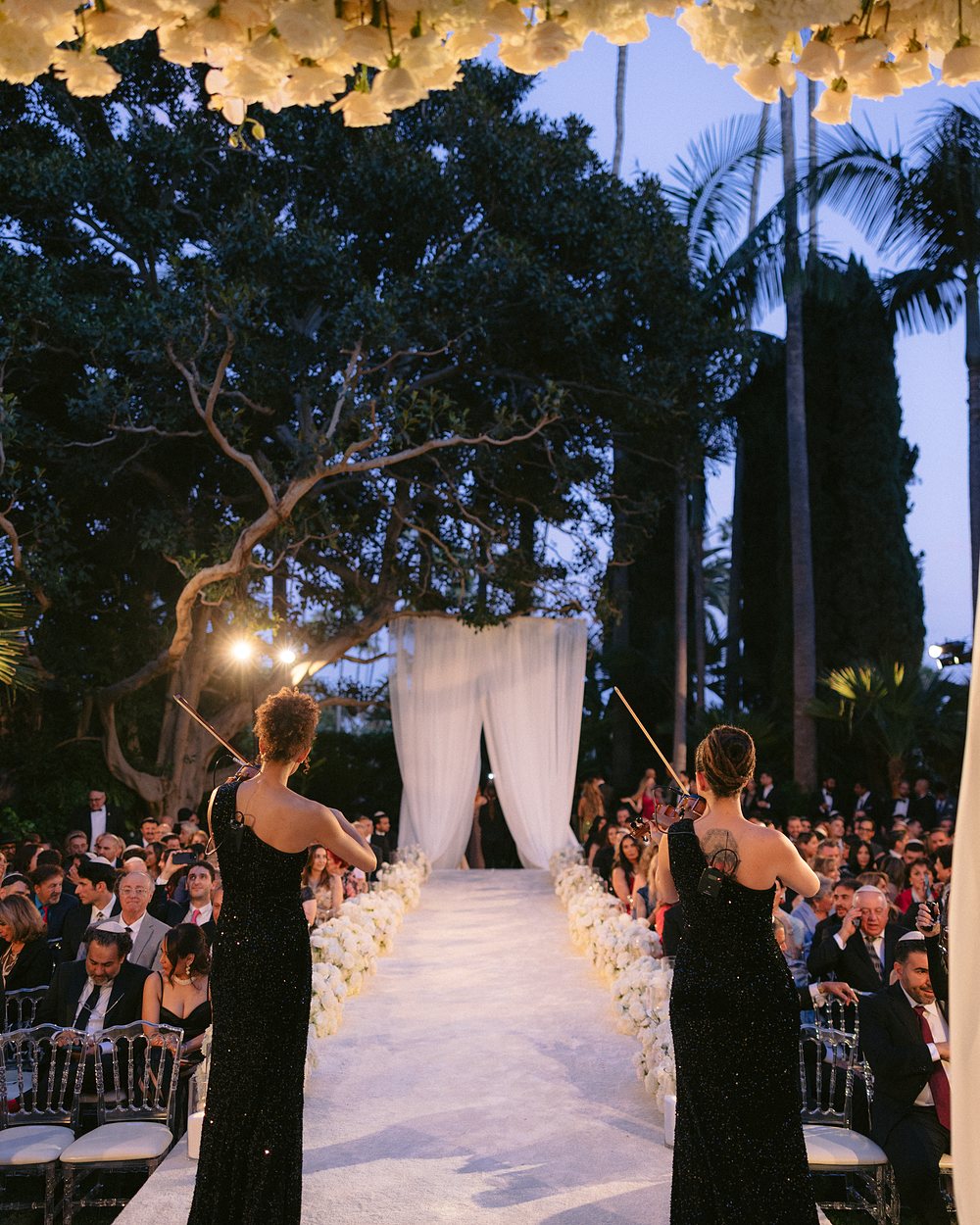 Two violinists playing at the head of an outdoor wedding aisle beneath a floral canopy