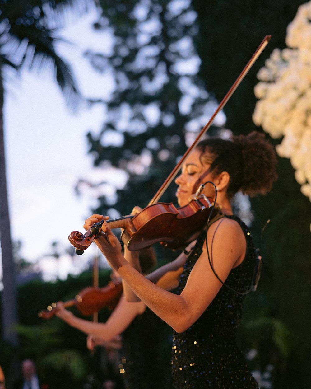 Violinist in a black sequined dress playing outdoors at dusk, a second violinist behind her