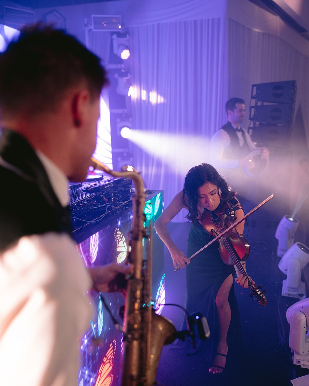 A violinist performing beside a lit DJ booth under stage lighting at a reception