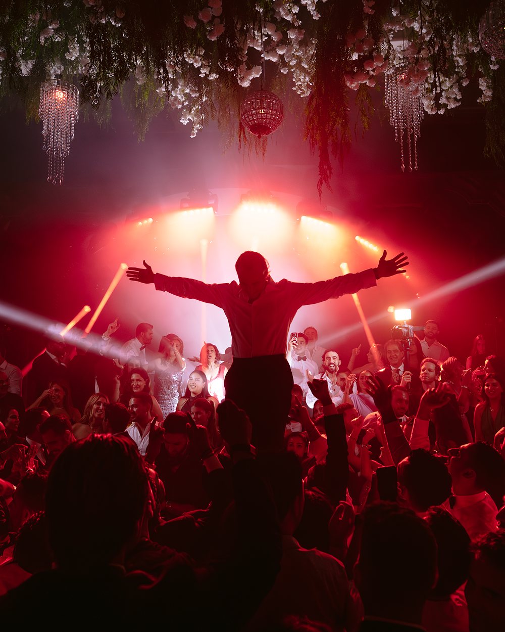 Guest lifted above the crowd with arms spread, silhouetted against red stage light under a floral ceiling